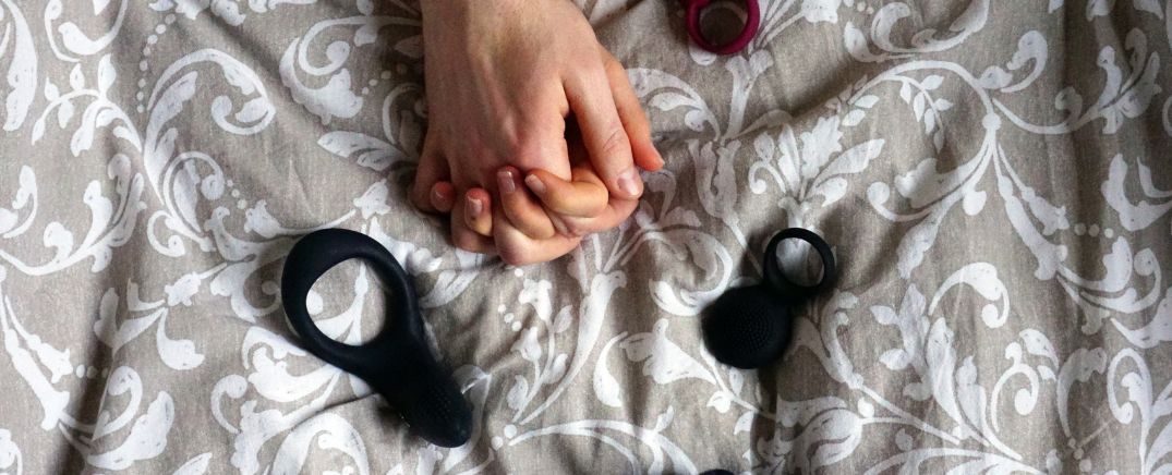 Dos manos entrelazadas sobre una cama junto a anillos sexuales, simbolizando la conexión y el deseo compartido.